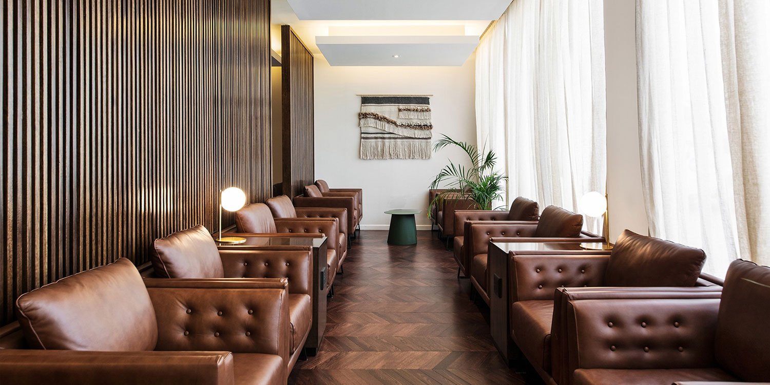 A very rich looking scene of the corner of the lounge features a walkway with large dark brown leather lounge chairs face each other on either side of the walls. at the end is a small round end table and some floor plants.