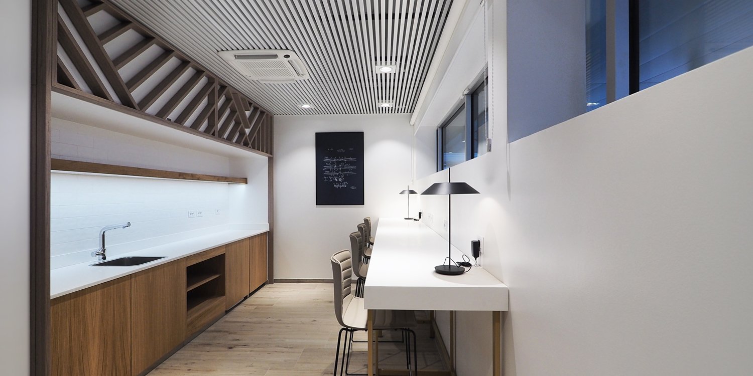 Work counter with four chairs and desk lamps opposite a timber kitchenette in the Aviapartner executive lounge at Rome Ciampino.