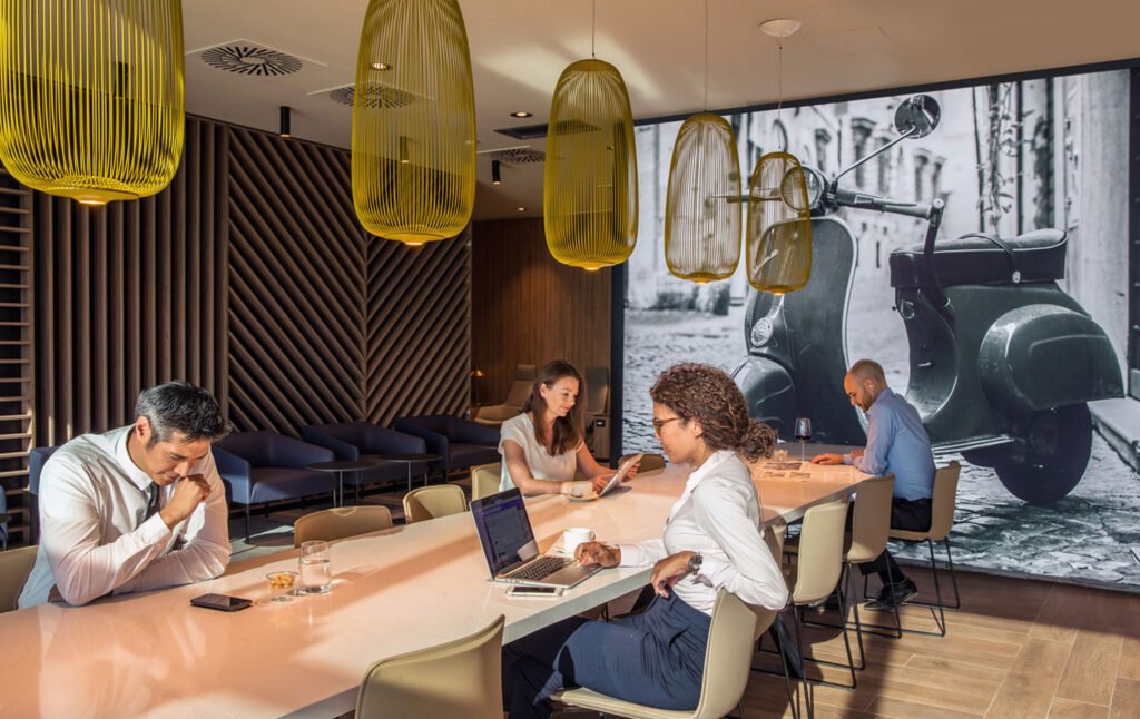 Guests working and dining at a communal table beneath yellow pendant lights beside a large Vespa photograph in the Star Alliance Lounge at Rome Fiumicino Airport.